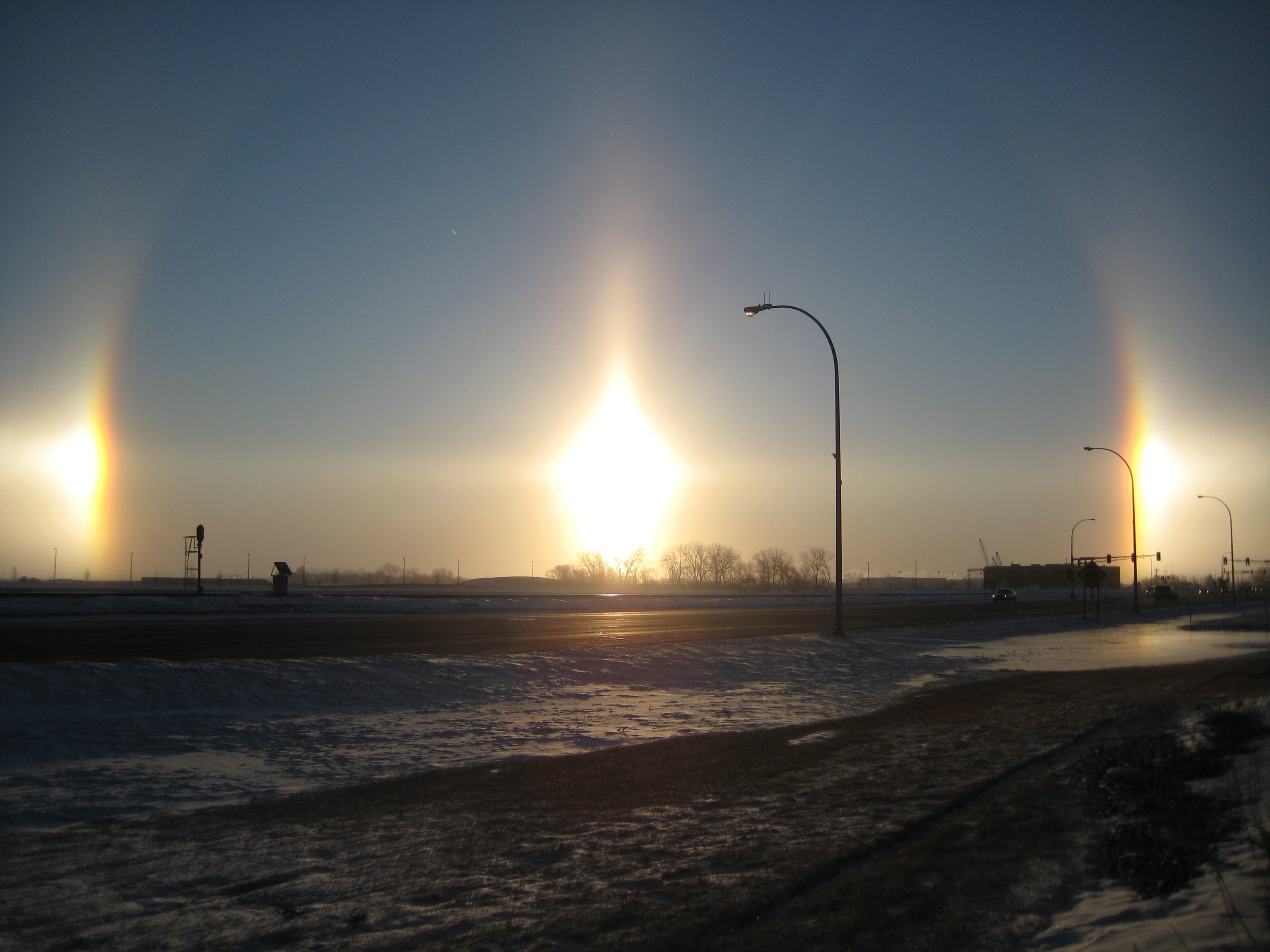 Modern colour photograph of a winter sky. The Sun sits low behind a bare tree, with two bright patches of light at the same height on either side of it, each showing faint colour, joined by a pale horizontal band.