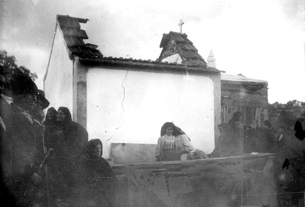 Black-and-white photograph of a small chapel with its roof destroyed and walls broken open, rubble on the ground, and a group of people standing beside the damage.