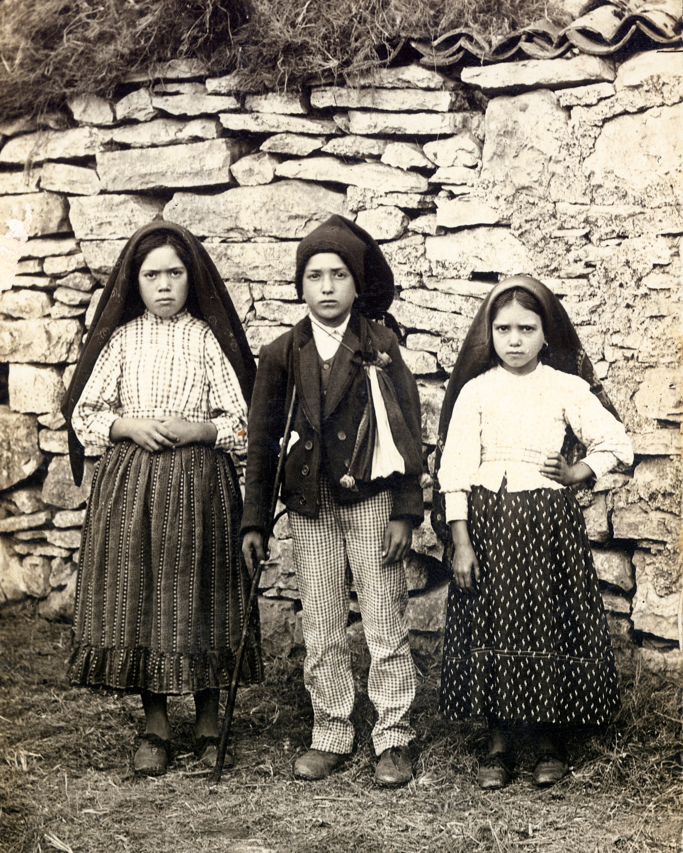 Black-and-white studio-style portrait of three children standing together outdoors. The two girls wear headscarves and long skirts; the boy stands between them in a jacket and hat, holding its brim.