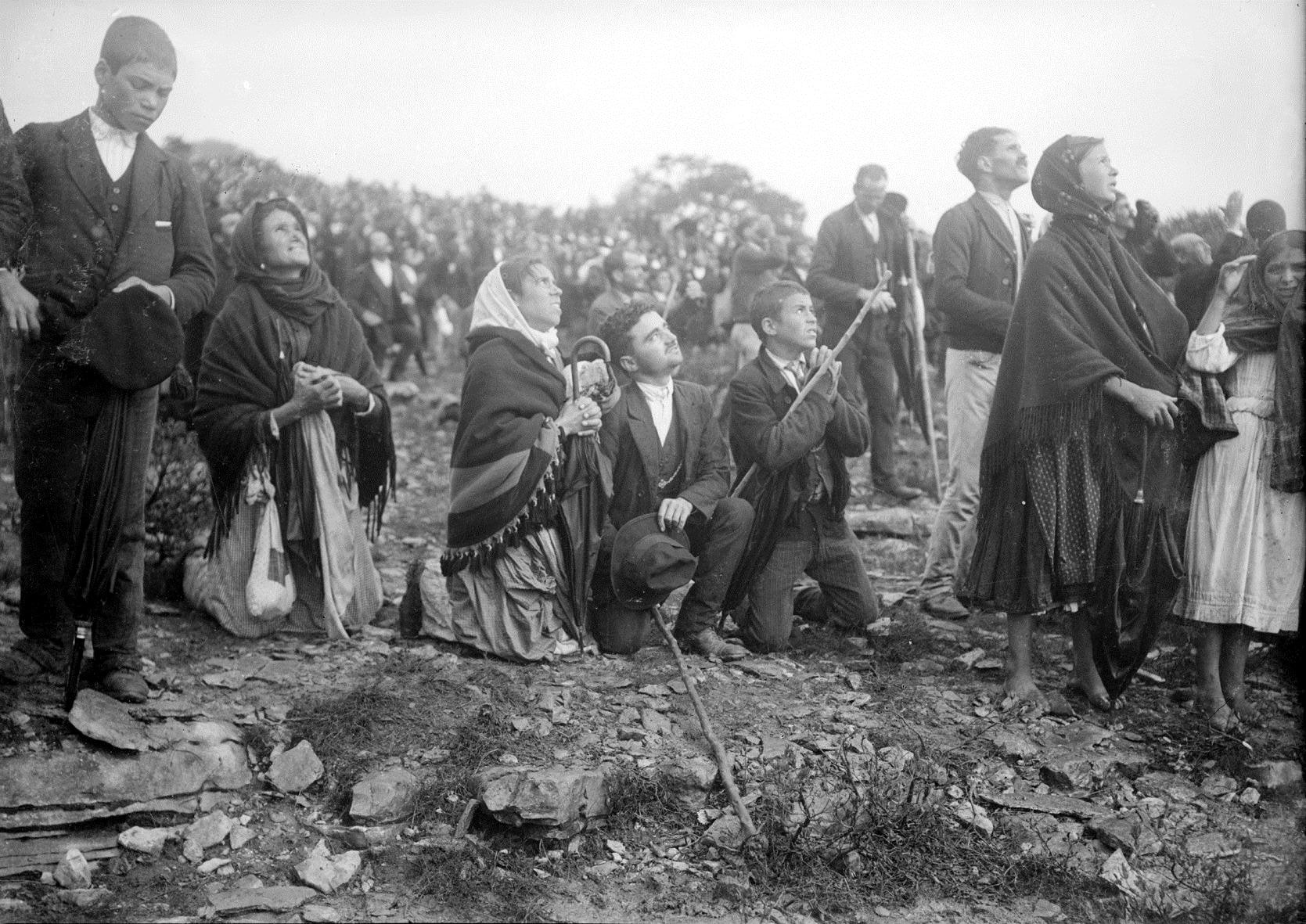 Black-and-white photograph of a large crowd at Cova da Iria in October 1917, many people looking upward. Figures are packed close together across the whole frame, most in hats and headscarves, faces tilted towards the sky.