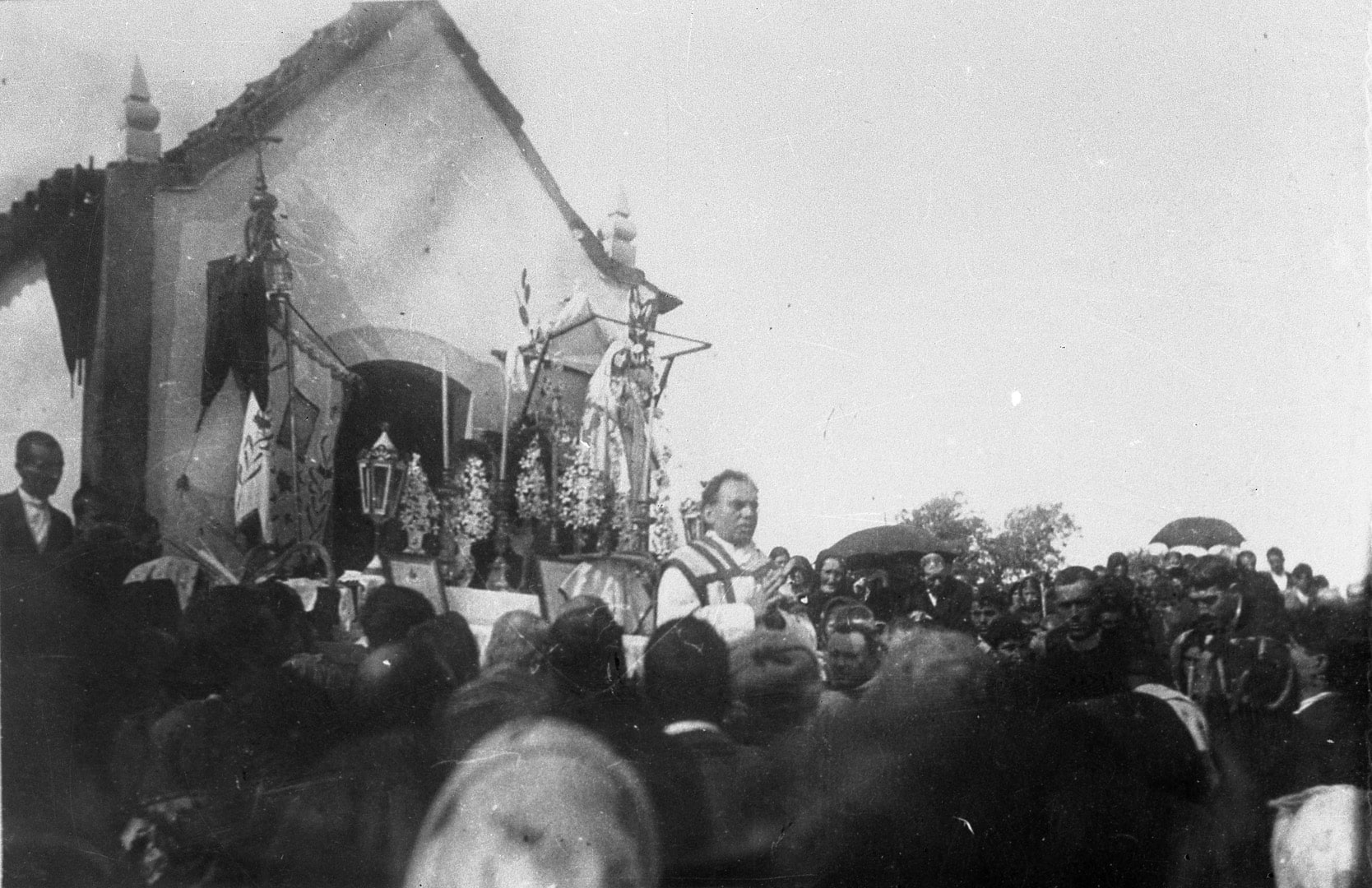 Black-and-white photograph of a crowd gathered on open ground around a very small single-room chapel, with a canopy erected beside it and figures standing on the roof line of the surrounding slope.