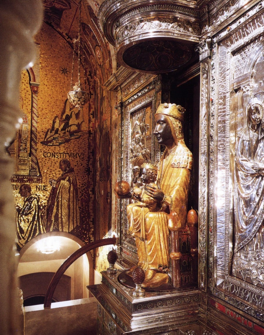 Seated Romanesque wooden Virgin and Child known as the Moreneta. The figure sits frontally with the child on her lap, one hand raised holding a sphere; the carved faces and hands are dark, the robes gilded, and the whole is set against a lit recess.