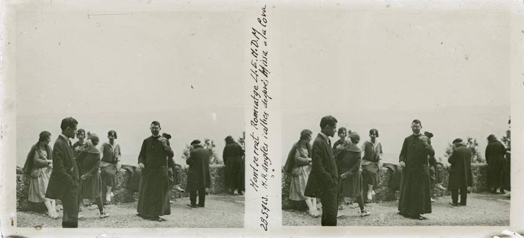 Wide black-and-white photograph of a large group of people standing closely packed on a terrace outside a chapel set against rock, most facing the camera, many in hats and coats.