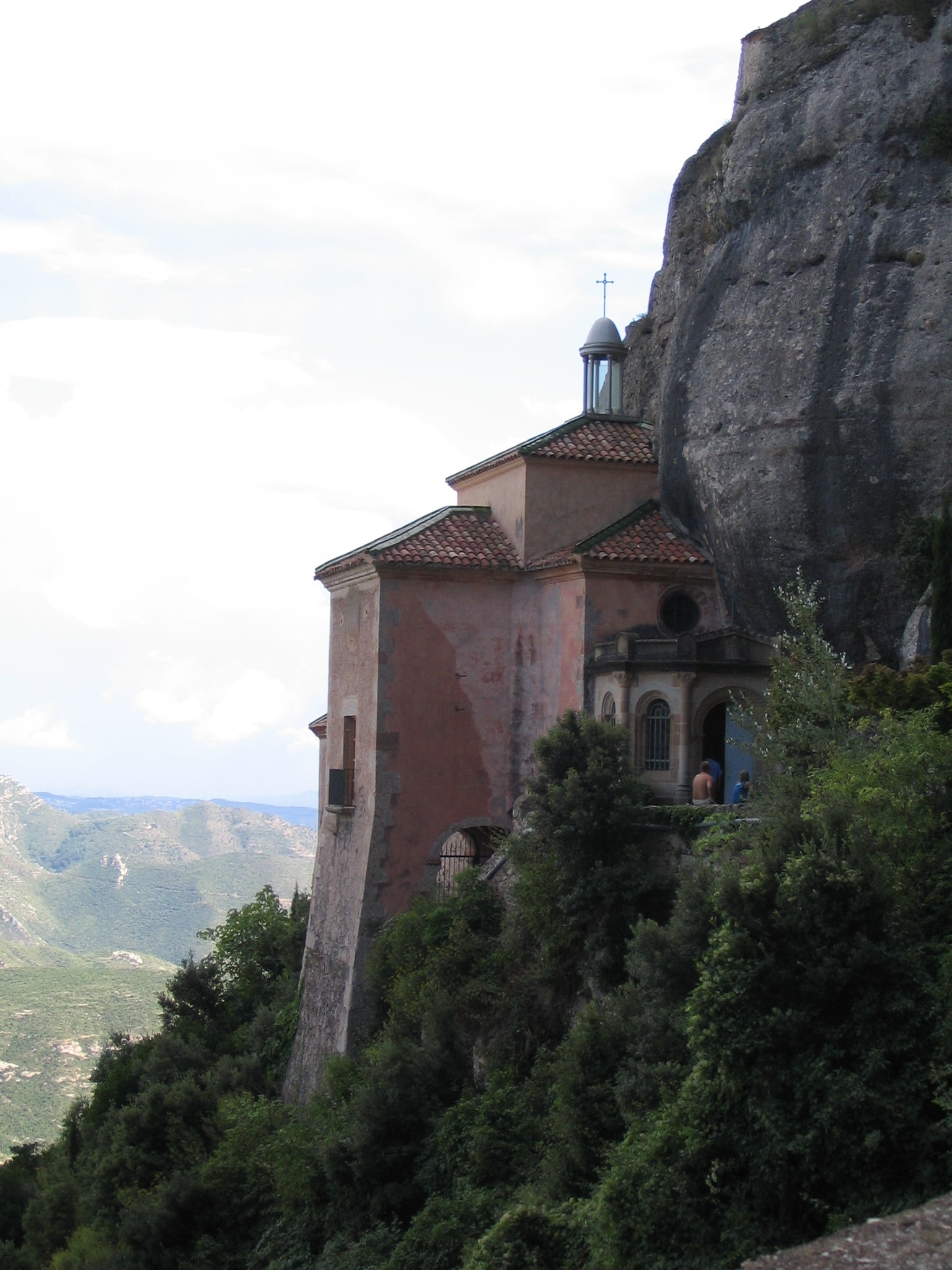 Photograph of the Santa Cova chapel built into the steep rock face at Montserrat. A small stone building with a low tower stands on a ledge against a wall of grey conglomerate rock, with wooded slopes below and bare pinnacles above.